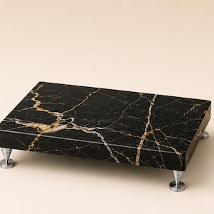 Puede incluir: Un soporte de exhibición rectangular de mármol negro con vetas blancas y doradas prominentes en toda la superficie. La losa está sostenida por cuatro pequeñas patas metálicas cónicas de tono plateado en las esquinas.
