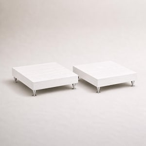 Puede incluir: Dos plataformas cuadradas con un acabado de veta de madera blanca, cada una apoyada sobre cuatro pequeños pies metálicos en forma de cono. Las plataformas están colocadas una al lado de la otra, mostrando una superficie plana y un diseño bajo.