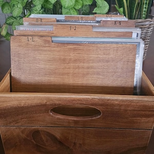 Puede incluir: Una caja de almacenamiento de discos de madera con asa, que contiene discos de vinilo. La caja tiene separadores etiquetados A-E, F-J, K-N, O-R, S-V y W-Z. Se aprecia la veta de la madera.