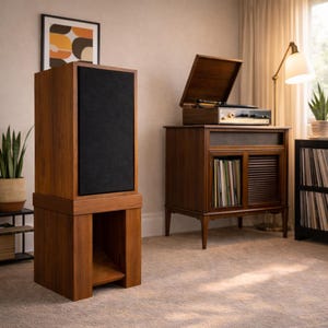 May include: A tall, wooden speaker with a black fabric front panel. The speaker sits atop a matching wooden base with an open shelf. A vintage record player and cabinet are in the background, suggesting a retro audio setup.