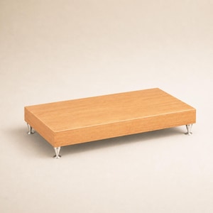 Puede incluir: Un expositor rectangular de madera con un acabado de veta de madera marrón claro. La plataforma está sostenida por cuatro pequeñas patas de metal de color plateado con forma cónica, que proporcionan una ligera elevación sobre la superficie.