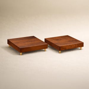 May include: Two square, wooden platforms with a rich, dark brown finish. Each platform has four small, gold-colored feet. The wood grain is visible, adding to the natural aesthetic. The platforms are set against a neutral background.