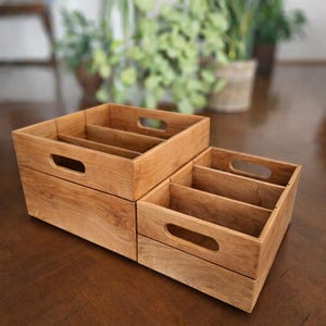 May include: Two wooden storage boxes with a natural finish. Each box has a rectangular shape with a handle cut out on the front. The boxes are divided into compartments, perfect for organizing items. The boxes are made of wood.