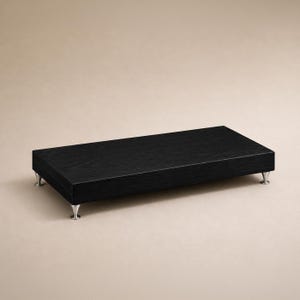 May include: A rectangular black wooden display riser with a visible wood grain texture. The platform is elevated by four small, metallic, cone-shaped feet positioned at each corner, providing a stable base for the flat, horizontal surface.