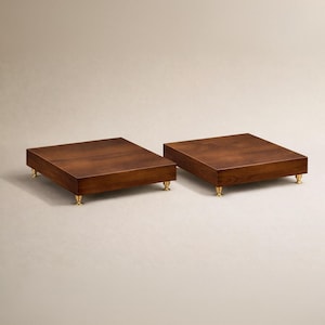 May include: Two square, wooden speaker stands with a rich, dark brown finish. Each stand is supported by four small, gold-colored feet. The stands are placed side-by-side against a neutral background, showcasing their simple, elegant design.