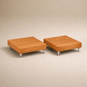 May include: Two wooden speaker stands with a square shape and a light brown finish. Each stand has four silver-coloured, cone-shaped feet. The stands are designed to elevate speakers.