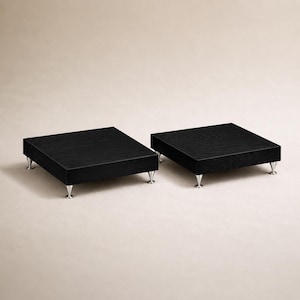 May include: Two square display risers featuring a black wood grain finish on the top surface. Each riser is supported by four small, silver-toned metal feet that are conical in shape, providing a slight elevation for the flat black platforms.