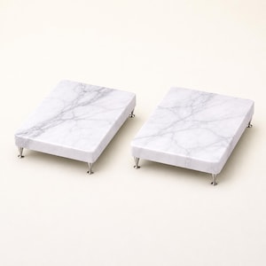 Puede incluir: Dos tablas rectangulares de mármol con vetas grises. Cada tabla tiene cuatro patas de metal plateado. Las tablas se muestran sobre un fondo blanco. Ideales para servir aperitivos o exhibir objetos pequeños.