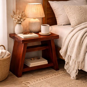 May include: A wooden nightstand with a lamp, books, and a mug of coffee. The lamp has a beige shade and a speckled base. A vase of dried flowers sits next to the lamp. A woven basket and a rug are in the foreground.