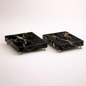 May include: Two rectangular black marble serving trays with gold veining. Each tray has four small, gold-colored feet. The trays are displayed on a white background, showcasing their elegant design.