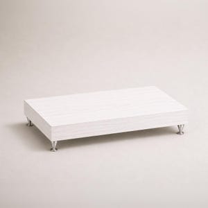 May include: A rectangular white wooden riser with a subtle wood grain texture. The platform is elevated by four small, metallic, cone-shaped feet positioned at each corner, providing a stable base for this display stand.