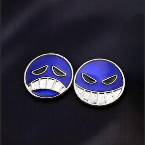 May include: Two circular, blue enamel pins with silver-coloured trim. One pin shows a sad face, the other a smiling face. The pins are set against a dark blue background.