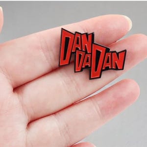 May include: A red and black enamel pin featuring the text "DAN VA DAN". The pin has a black border and is held in a hand. The text is in a stylized font.