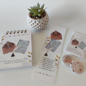 Pode incluir: Um conjunto de artigos de papelaria com tema literário. Inclui um caderno espiral, um marcador, um cartão pequeno e dois pinos redondos. Cada item apresenta uma ilustração de envelope, livro e pena, com texto em português.