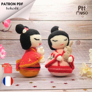 May include: Two crocheted Kokeshi dolls, one in red and orange and the other in pink and white, with black hair and closed eyes. The doll on the right is holding a small crocheted rabbit.