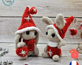 May include: Two crocheted bunny figurines wearing red Santa hats and scarves. One bunny holds a red lantern with a white star design. The bunnies are gray and white, with black eyes, and are set against a wooden background.