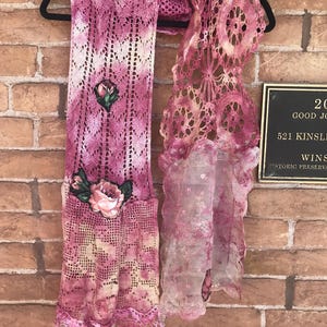 Könnte beinhalten: Handgefertigter rosa-weißer Häkelschal mit floralen Verzierungen. Der Schal hat ein detailliertes Lochmuster und ist mit gestickten Rosen verziert. Der Schal hängt an einem schwarzen Kleiderbügel vor einer Backsteinmauer.