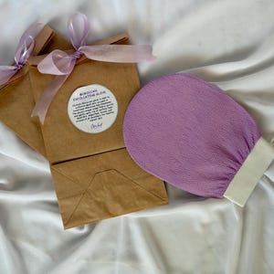 Può includere: Un guanto esfoliante viola con polsino bianco è appoggiato accanto a due sacchetti di carta marrone legati con nastri lavanda. Le borse hanno un'etichetta circolare che dice "Moroccan Exfoliating Glove". Lo sfondo è un tessuto bianco e serico.