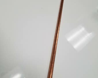 May include: A single, straight copper rod, lying diagonally against a white surface. The rod has a smooth, cylindrical shape and a warm, reddish-brown color. The lighting creates a subtle highlight along its length.