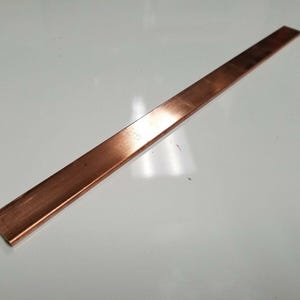 Può includere: Una lunga barra rettangolare di rame con una superficie lucida. La barra è di colore marrone rossiccio e sembra essere fatta di metallo solido. La barra è dritta e ha una larghezza e uno spessore costanti. La barra di rame viene probabilmente utilizzata in edilizia o in applicazioni elettriche.