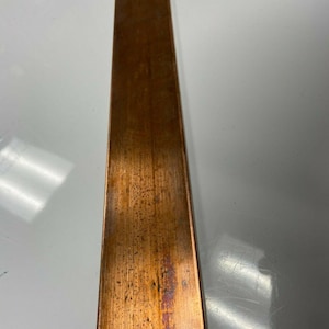 May include: A long, rectangular strip of copper metal with a flat surface. The material displays a warm, reddish-brown metallic tone with visible natural oxidation, slight surface variations, and a smooth, straight-edged finish throughout.