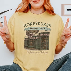May include: A yellow t-shirt with a graphic of Honeydukes Sweets Shop from the Harry Potter series. The graphic features a storefront with the words 'Honeydukes Sweets Shop' above the entrance. The text 'Hogsmeade Village' is below the storefront. The t-shirt is on a person wearing blue jeans.