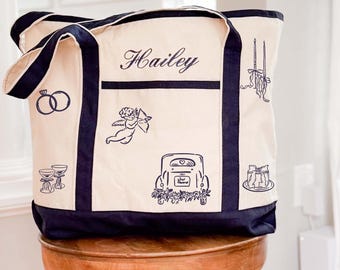 May include: A cream-colored canvas tote bag with navy blue trim and handles. The bag features the name Hailey embroidered in navy blue script, along with embroidered navy blue icons including wedding rings, a cupid, a car, and champagne flutes.