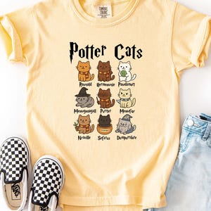 May include: A light yellow t-shirt with the text "Potter Cats" and illustrations of cats dressed as characters. The shirt is paired with denim shorts and black and white checkered slip-on shoes.