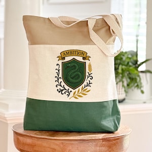 May include: A canvas tote bag with a tan top, cream middle, and green bottom. The cream section features a crest with a green snake, the word "AMBITION", and decorative elements. The bag has cream-colored handles.
