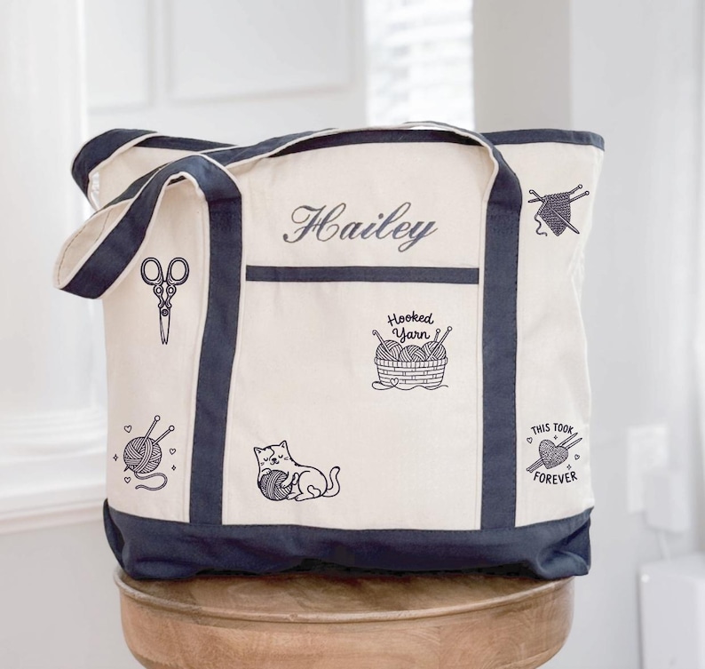 May include: A cream-coloured canvas tote bag with dark navy blue trim and handles. The bag features black embroidered designs including scissors, a cat playing with yarn, a basket labelled Hooked Yarn, and text reading Hailey and This Took Forever.