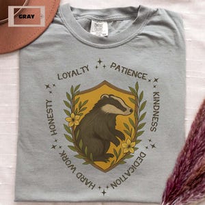 Puede incluir: Camiseta gris con un dise&ntilde;o de escudo de tej&oacute;n. El escudo presenta una ilustraci&oacute;n de tej&oacute;n dentro de un escudo dorado, rodeado por el texto "Lealtad, Paciencia, Honestidad, Trabajo duro, Dedicaci&oacute;n, Amabilidad".
