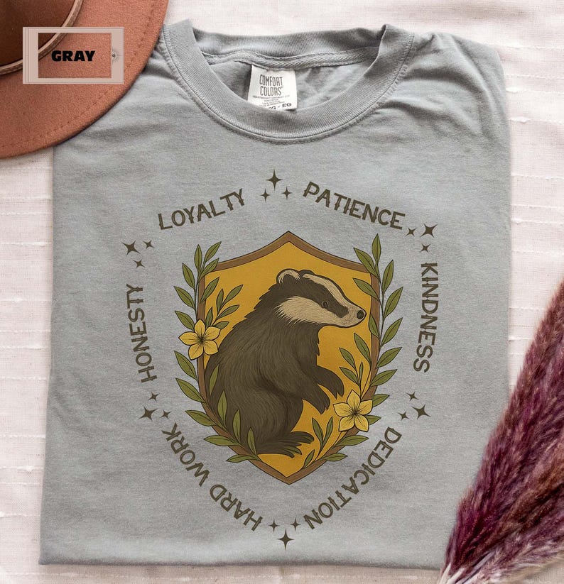 Puede incluir: Camiseta gris con un dise&ntilde;o de escudo de tej&oacute;n. El escudo presenta una ilustraci&oacute;n de tej&oacute;n dentro de un escudo dorado, rodeado por el texto "Lealtad, Paciencia, Honestidad, Trabajo duro, Dedicaci&oacute;n, Amabilidad".