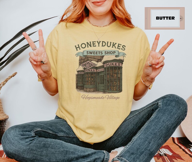 May include: A yellow t-shirt with a graphic of Honeydukes Sweets Shop from the Harry Potter series. The graphic features a storefront with the words 'Honeydukes Sweets Shop' above the entrance. The text 'Hogsmeade Village' is below the storefront. The t-shirt is on a person wearing blue jeans.