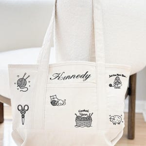 May include: A cream-coloured canvas tote bag featuring black line art illustrations of a yarn ball with needles, scissors, a cat playing with yarn, a basket of yarn labelled Hooked Yarn, a sheep, and a person knitting with the text Just One More Row... I swear. The name Kennedy is embroidered in black script.
