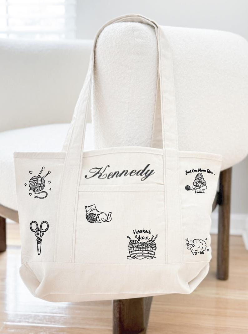 May include: A cream-coloured canvas tote bag featuring black line art illustrations of a yarn ball with needles, scissors, a cat playing with yarn, a basket of yarn labelled Hooked Yarn, a sheep, and a person knitting with the text Just One More Row... I swear. The name Kennedy is embroidered in black script.