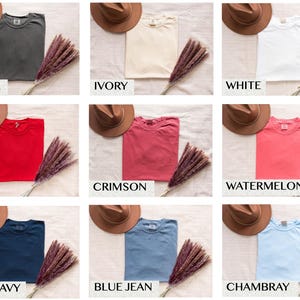 May include: A flat lay of ten different colored t-shirts with a hat on top of each shirt. The colors are pepper, ivory, white, red, crimson, watermelon, true navy, blue jean, and Chambray.
