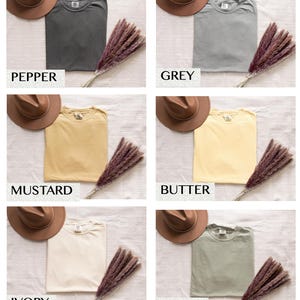 Puede incluir: Colecci&oacute;n de seis camisetas plegadas en varios colores: pimienta, gris, mostaza, mantequilla, marfil y caqui. Cada camiseta se combina con un sombrero marr&oacute;n y un manojo de hierba seca de color p&uacute;rpura-marr&oacute;n. El color de cada camiseta est&aacute; etiquetado.