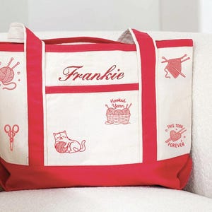 May include: A cream-coloured canvas tote bag with red trim and handles. The bag features red embroidered designs including a ball of yarn, scissors, a cat playing with yarn, a basket labelled Hooked Yarn, and a heart with the text This Took Forever.