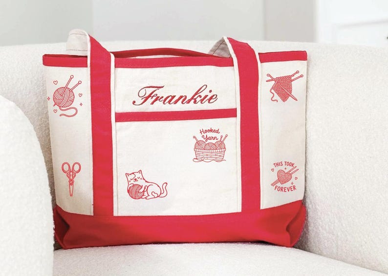May include: A cream-coloured canvas tote bag with red trim and handles. The bag features red embroidered designs including a ball of yarn, scissors, a cat playing with yarn, a basket labelled Hooked Yarn, and a heart with the text This Took Forever.
