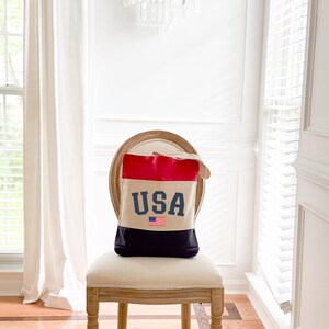 May include: A canvas tote bag with a patriotic design. The top section is red, the middle is beige, and the bottom is black. The word "USA" is printed in blue, with a small American flag below.