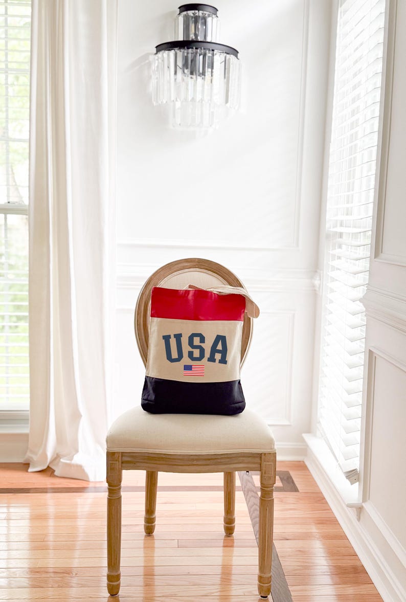 May include: A canvas tote bag with a patriotic design. The top section is red, the middle is beige, and the bottom is black. The word "USA" is printed in blue, with a small American flag below.