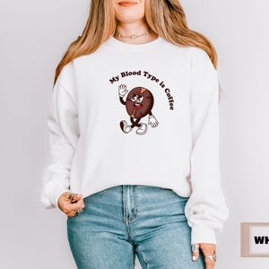 White sweatshirt featuring a playful design with a smiling cartoon coffee bean character waving and walking. Above the illustration, bold curved text reads 'My Blood Type is Coffee' in dark brown.