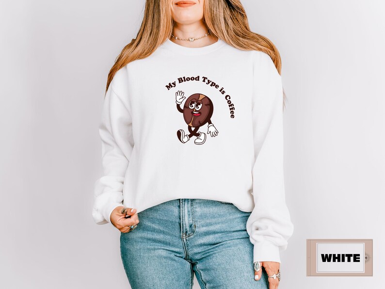White sweatshirt featuring a playful design with a smiling cartoon coffee bean character waving and walking. Above the illustration, bold curved text reads 'My Blood Type is Coffee' in dark brown.