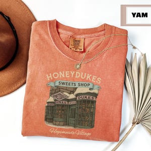 Op de afbeelding: Een perzikkleurig T-shirt met een afbeelding van Honeydukes Sweets Shop uit de Harry Potter-serie. De afbeelding toont een winkel met sneeuw op het dak en de tekst "Honeydukes Sweets Shop" en "Hogsmeade Village".