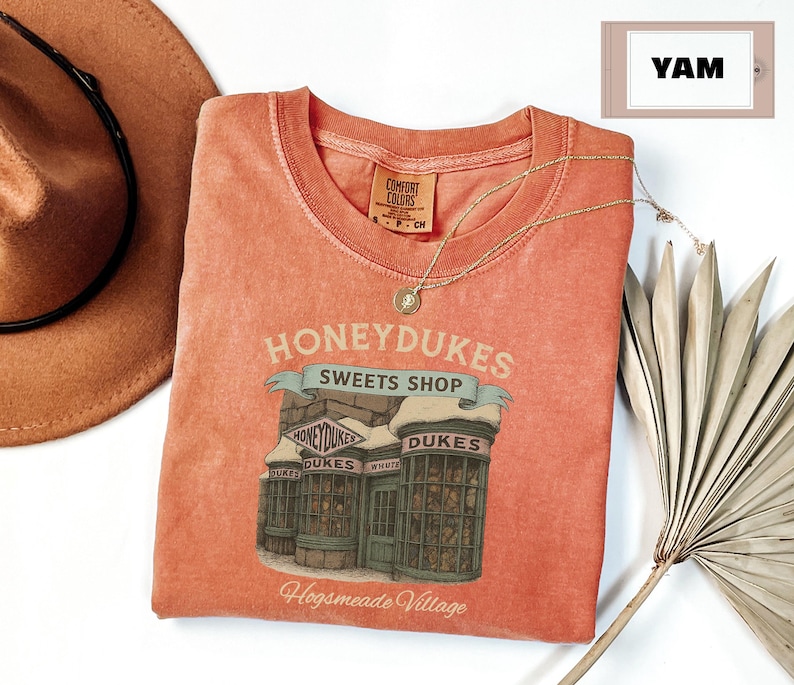 Op de afbeelding: Een perzikkleurig T-shirt met een afbeelding van Honeydukes Sweets Shop uit de Harry Potter-serie. De afbeelding toont een winkel met sneeuw op het dak en de tekst "Honeydukes Sweets Shop" en "Hogsmeade Village".