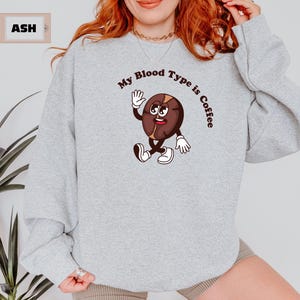 Gray sweatshirt featuring a playful design with a smiling cartoon coffee bean character waving and walking. Above the illustration, bold curved text reads 'My Blood Type is Coffee' in dark brown.