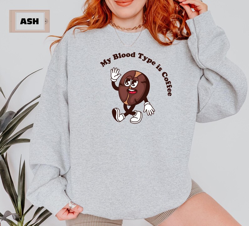 Gray sweatshirt featuring a playful design with a smiling cartoon coffee bean character waving and walking. Above the illustration, bold curved text reads 'My Blood Type is Coffee' in dark brown.