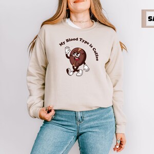 Beige sweatshirt featuring a playful design with a smiling cartoon coffee bean character waving and walking. Above the illustration, bold curved text reads 'My Blood Type is Coffee' in dark brown.