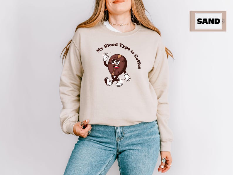 Beige sweatshirt featuring a playful design with a smiling cartoon coffee bean character waving and walking. Above the illustration, bold curved text reads 'My Blood Type is Coffee' in dark brown.