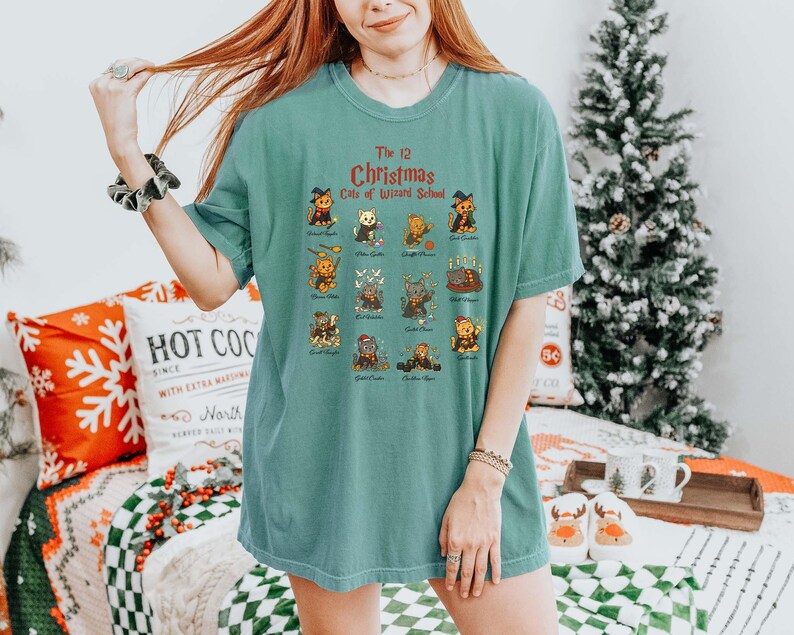 Woman wearing a light green oversized t-shirt featuring a holiday design titled “The 12 Christmas Cats of Wizard School,” showing cute illustrated cats dressed as magical characters. Festive Christmas decor in the background including tree, pillows, and seasonal accents.